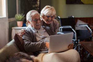 Älteres Paar mit Laptop auf dem Sofa, Rollstuhl im Hintergrund.