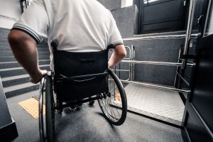 Rollstuhlfahrer vor einer Treppe und einem Hublift am Gebäudeeingang.