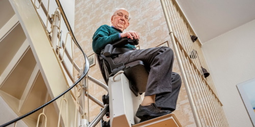 Älterer Mann fährt mit einem Treppenlift die Treppe hinunter.